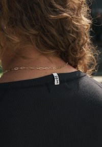 Zwart geribd kledingstuk met een ronde hals, met een wit label met de tekst "self." Een dunne gouden ketting is zichtbaar rond de nek.