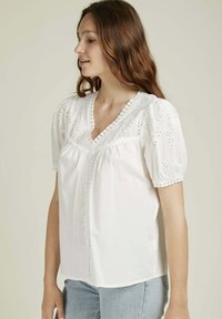 Blouse en coton blanc avec des détails brodés et une finition en dentelle aux manches et au col. Manches courtes à volants et fermeture à boutons à l'avant.