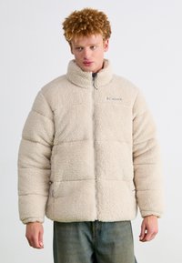 Beige fleece jacket with a high collar, zip closure, and horizontal quilted pattern, featuring side pockets and a logo on the chest.