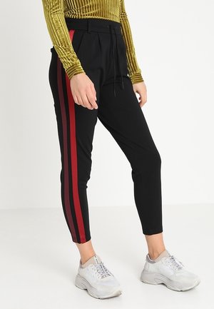 Person iført sorte tilspidsede bukser med to røde lodrette striber på siden, hvide chunky sneakers og en langærmet gylden tekstureret top.