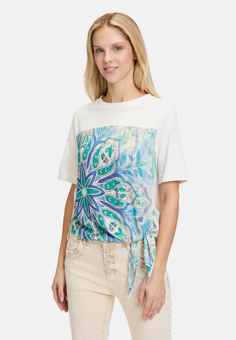 Hvid kortærmet T-shirt med en farverig, mønstret front, der har blå og grønne blomsterdesigns; inkluderer detalje med knude i siden.