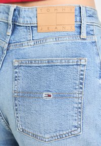 Šviesiai mėlyni džinsiniai šortai su odiniu žymeniu, pažymėtu "TOMMY JEANS." Užpakalinėje kišenėje yra mažas raudonas, baltas ir mėlynas logotipas.