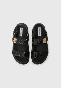 Sandales noires à glissière avec des sangles en simili cuir matelassé, des accents en métal doré et une semelle intérieure lisse et rembourrée. Nom de la marque visible à l'intérieur.
