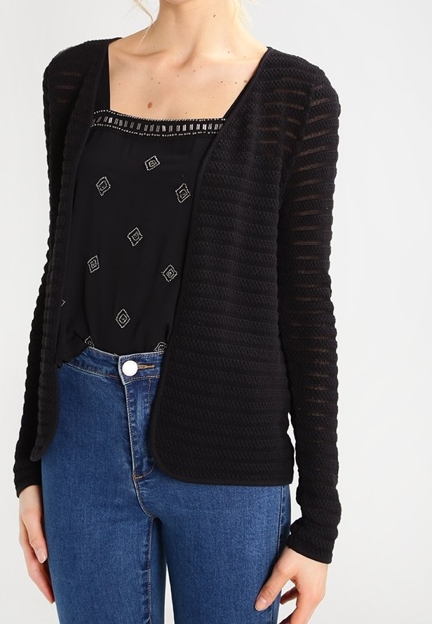 Black cardigan with a textured knit design, open front, long sleeves, and a relaxed fit, worn over a black patterned blouse and blue jeans.