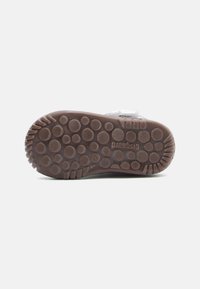 Suela de zapato de goma marrón con patrones de agarre circulares y prominencias, que presenta la marca "Gispu". La parte superior tiene un textil suave gris con correa.