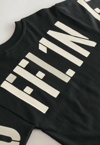 Sort T-shirt med store hvide blokbogstaver, med rund hals og korte ærmer. Stoffets tekstur ser blød og glat ud.