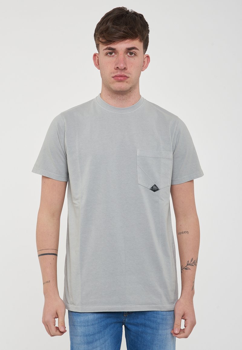 T-shirt grigio chiaro a maniche corte in cotone, con taschino sul petto e una toppa nera triangolare. Texture liscia, scollo a girocollo.