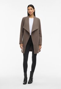 Abrigo marrón de mezcla de lana con un diseño de cuello abierto, combinado con una blusa blanca, jeans negros ajustados y botines negros.