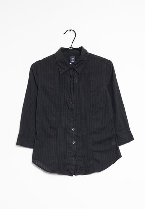 Camisa negra de manga larga con botones, con sutiles rayas verticales, cuello y detalles plisados en la parte delantera, colgada en una percha negra.