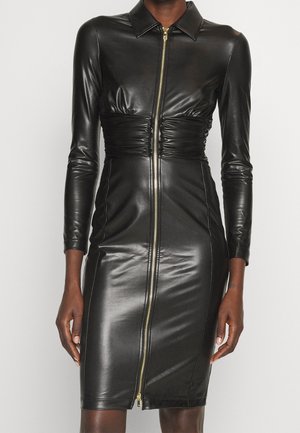 Femme portant une robe noire en similicuir moulante, à manches longues, taille froncée, col, et une fermeture éclair dorée pleine longueur à l'avant.