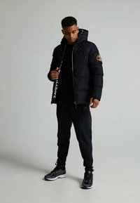 Black puffer jacket with a zip closure, hood, and logo patch. Paired with matching black pants and grey sneakers. Simple design, warm look.