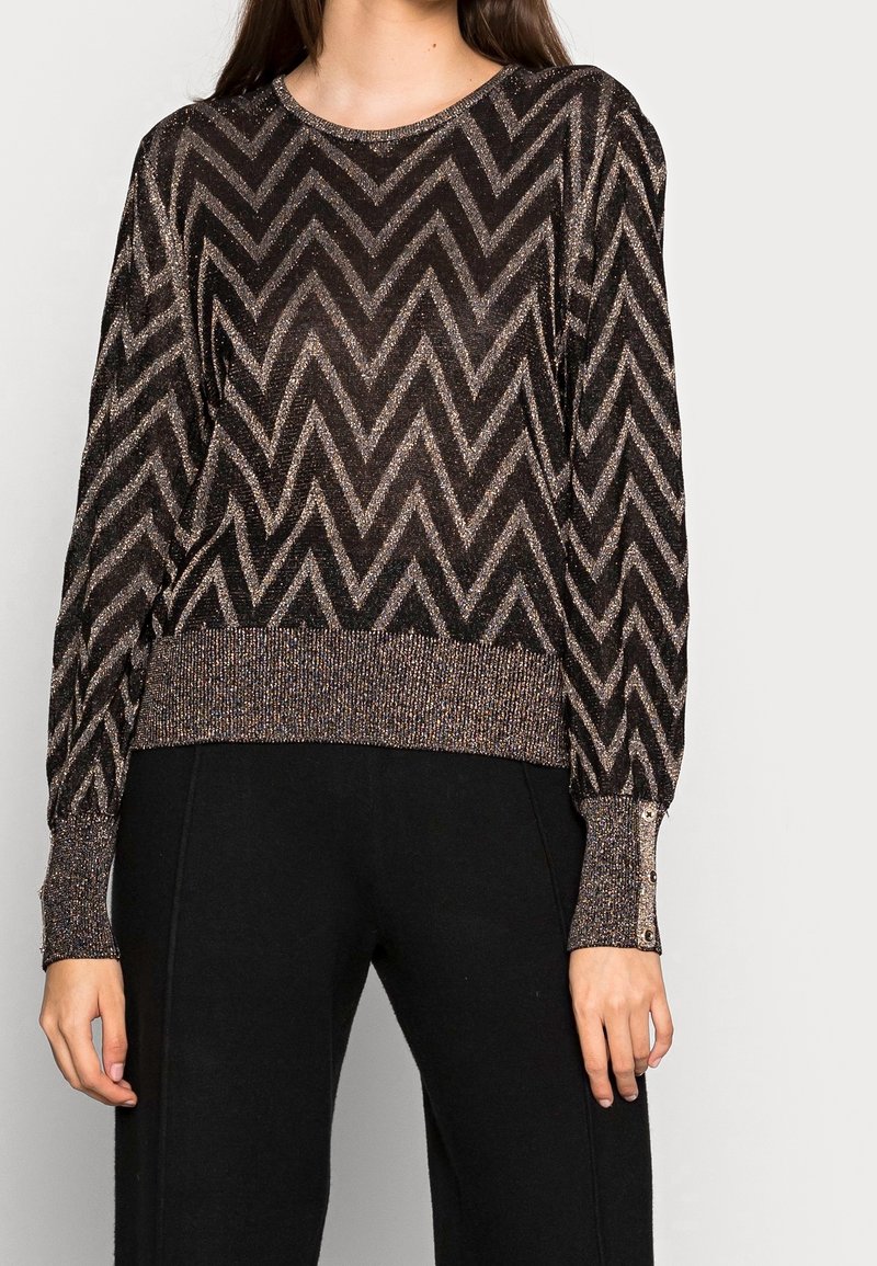Femme portant un pull à manches longues noir et bronze métallisé avec un motif chevron, des poignets boutonnés et un pantalon noir.