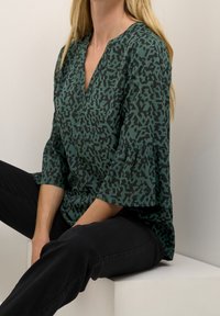 Blouse verte avec motif abstrait noir, encolure en V et manches évasées ; associée à un pantalon noir ; texture lisse, coupe décontractée.