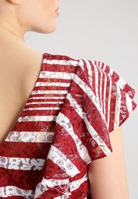 Blouse rayée rouge et blanche avec détails en dentelle, présentant des panneaux transparents et des manches courtes volantées. La vue de dos met en valeur le design de l'encolure.