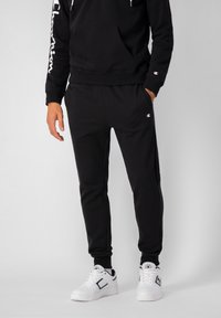 Sudadera de algodón negra con un bolsillo frontal y el logo blanco "Champion" en la manga. Combinada con joggers negros a juego y zapatillas blancas.
