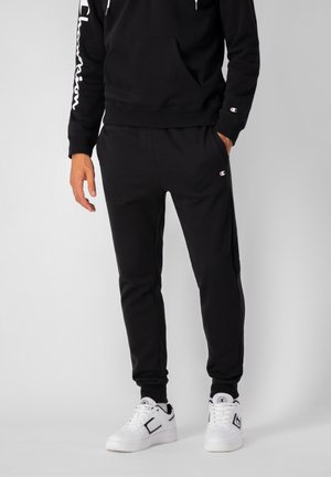 Felpa nera in cotone con tasca frontale e logo "Champion" bianco sulla manica. Abbinata a pantaloni della tuta neri e sneakers bianche.