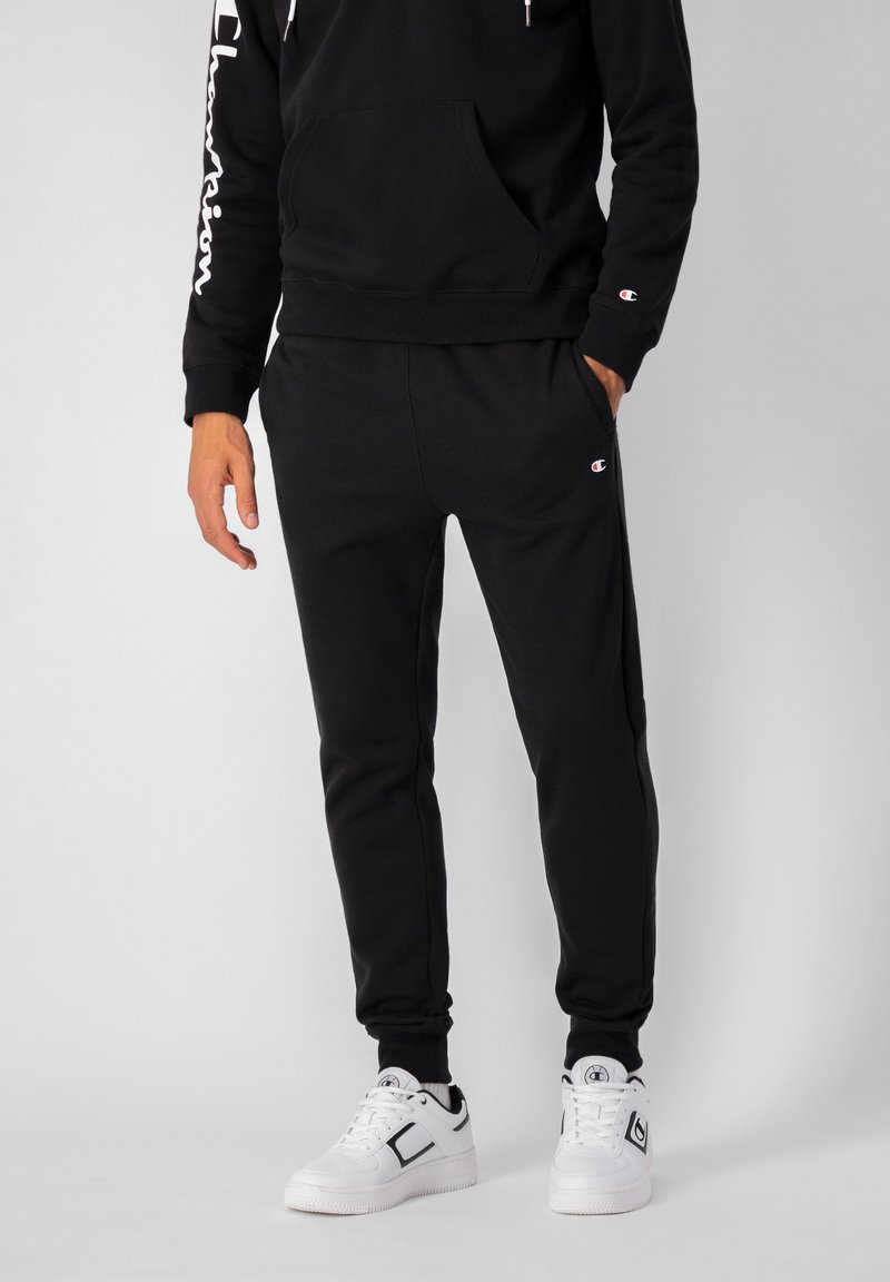 Sudadera de algodón negra con un bolsillo frontal y el logo blanco "Champion" en la manga. Combinada con joggers negros a juego y zapatillas blancas.