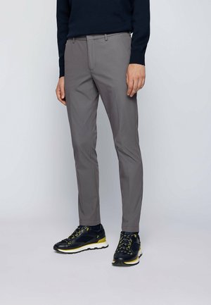 Man met grijze slim-fit broek, zwarte en gele sneakers en een donker topje met lange mouwen, staand tegen een effen lichte achtergrond.