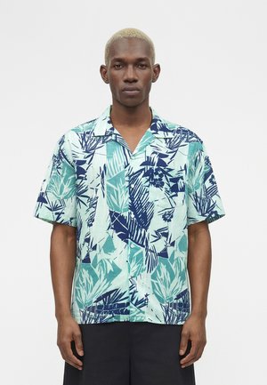 Mann iført kortermet knappeskjorte med blågrønt tropisk løvmønster, stående foran en ensfarget hvit bakgrunn.