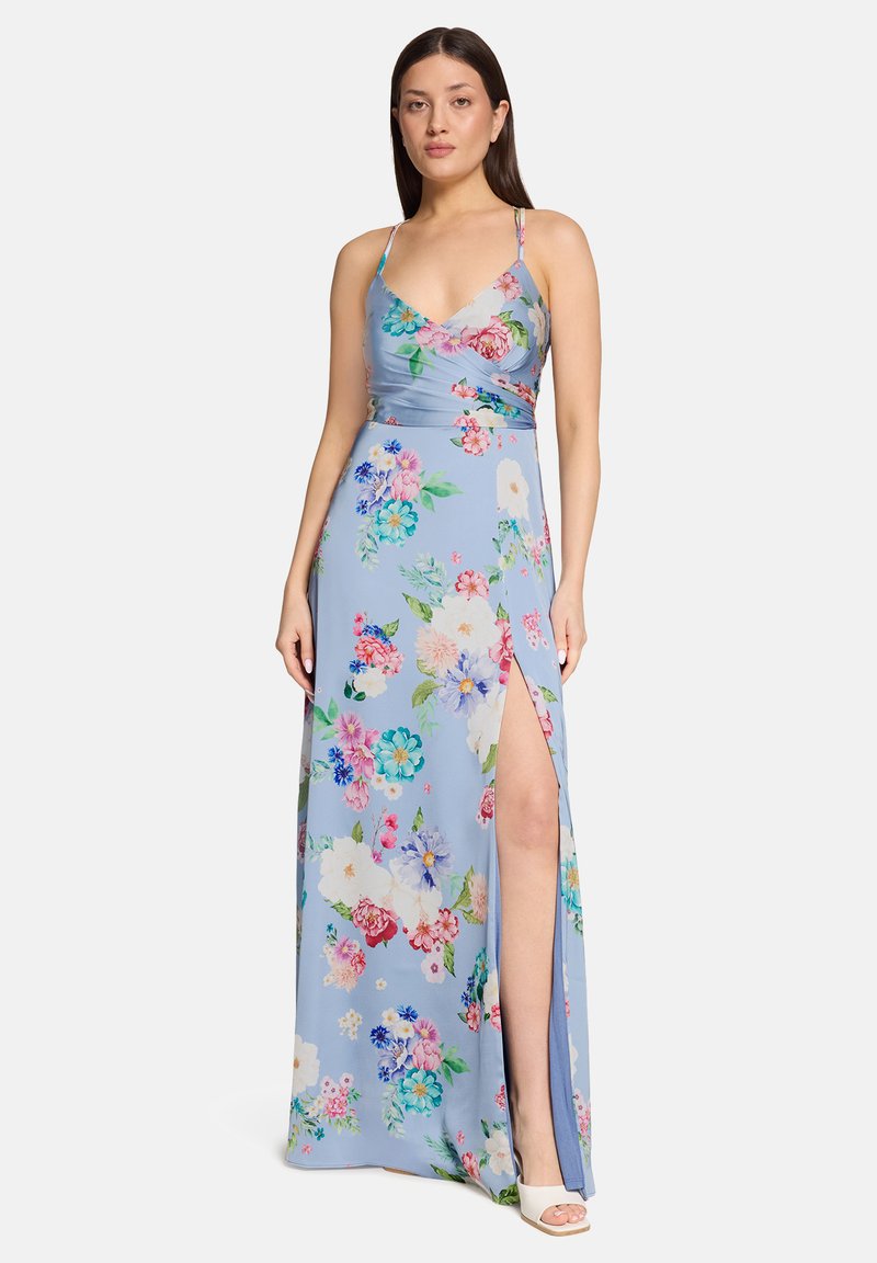 Femme portant une robe maxi fleurie bleu clair à fines bretelles et fendue sur le côté, associée à des talons ouverts blancs, debout devant un fond blanc.