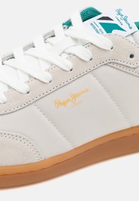 Zapatilla blanca y gris con acentos de ante, que presenta una suela de goma texturizada, cordones blancos y una etiqueta azul-verde que muestra el nombre de la marca.