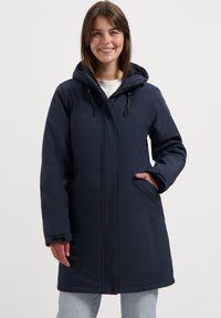 Vrouw die glimlacht, gekleed in een marineblauwe winterjas met capuchon, rits en zakken, in combinatie met lichtblauwe jeans, staand voor een effen achtergrond.