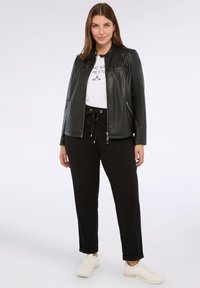 Kvinna stående iförd svart skinnjacka, vit t-shirt med grafik, svarta byxor med snörning och vita sneakers mot vit bakgrund.