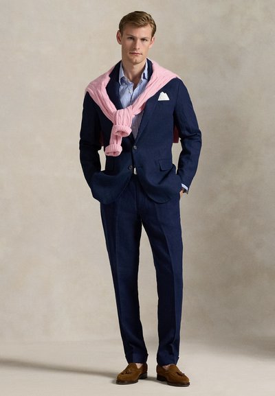 Marineblå dress med tettsittende design, lys blåstripet skjorte, rosa strikket genser lagt over, og brune loafers. Hvit lommetørkle-detalj.