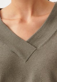 Pull vert olive avec un col en V. Confectionné dans un matériau doux et texturé. Présente un col côtelé et une coupe décontractée autour du col.