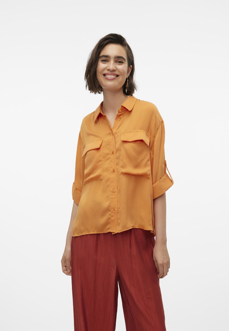 Camicia arancione con bottoni, dal taglio rilassato, con due tasche sul petto e maniche arrotolate, abbinata a pantaloni rossi a pieghe. Texture morbida.