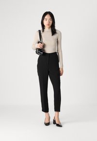 Top de manga larga beige con acanalado, pantalones negros ajustados y tacones de charol negros, sosteniendo un pequeño bolso negro de hombro. Diseño minimalista.