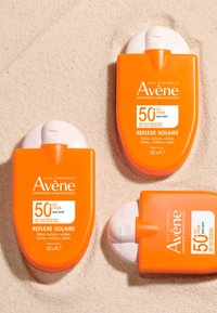 Three orange sunscreen bottles from Avène, labeled SPF 50+, with white accents, lying on beige sand. Each bottle contains 30 ml.