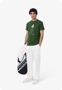 Vihreä puuvillapaita, jossa on graafinen kuva tikkaista ja tennispalloista. Yhdistettynä valkoisiin housuihin ja kannettuna tummansinistä tennisreppua.