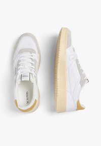 Zapatillas blancas con acentos de ante en gris claro, talón en contraste color tan, suela de goma texturizada y área de puntera perforada para transpirabilidad.