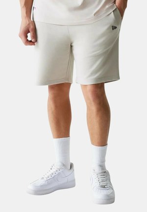 Personne portant un short beige clair avec des poches latérales, des chaussettes blanches côtelées mi-mollet et des baskets basses blanches, debout les mains dans les poches.