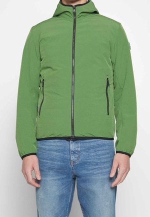 Mężczyzna w zielonej kurtce z kapturem na zamek błyskawiczny z czarnymi detalami zamka i niebieskich dżinsach na jednolitym białym tle.