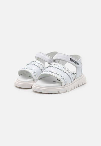 White sandals with a textured design, multiple straps, and Velcro closures. Notable logo detailing and a chunky white sole.