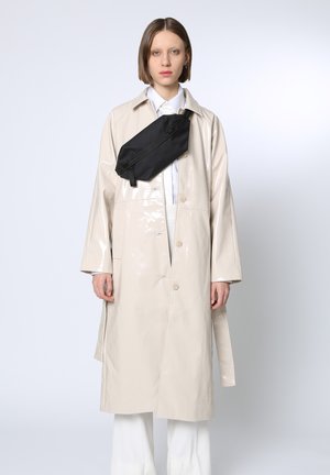 Beige glänzender PVC-Trenchcoat mit weiten Ärmeln, Vorderknöpfen und einer diagonalen schwarzen Gürteltasche über dem Körper getragen.