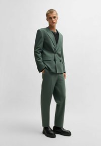 Grüner doppelt geknöpfter Anzug mit slim fit, passender Hose und einem schwarzen strukturierten Oberteil darunter. Schwarze Schuhe runden den Look ab.