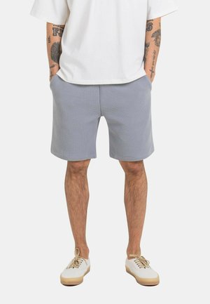 Man wearing light gray shorts, white short-sleeve shirt, white sneakers with beige soles, and hands in pockets, shown from waist down.