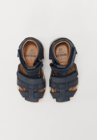 Mørkeblå lærsandaler med krydsede stropper, åbne tæer og justerbare Velcro-lukninger. Lysebrune indlægssåler med synlig mærkelogo.