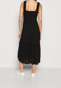 Vestido negro de longitud media con tirantes con volantes, diseño en capas y tela suave y ligera. Combinado con zapatillas blancas en la parte inferior.