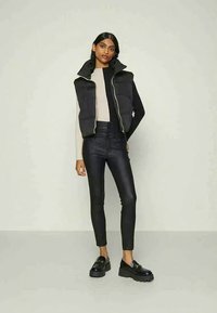 Μαύρο γιλέκο με υψηλό γιακά, συνδυασμένο με μπεζ μακρυμάνικο μπλουζάκι και μαύρες στενές τζιν. Διαθέτει σκούρα παπούτσια με χοντρή σόλα.