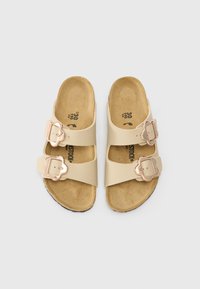 Beige sandaler med dubbel justerbara remmar med metallspännen. Texturerad fotbädd och ljus, flexibel sula.