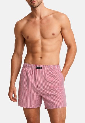 Oberkörperfreier Mann mit durchtrainiertem Oberkörper trägt pink-weiße vertikal gestreifte Boxershorts vor hellem, einfarbigem Hintergrund.