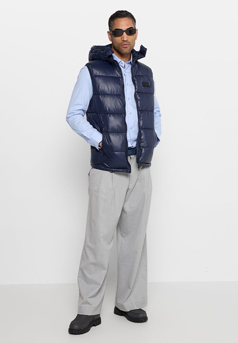 Veste matelassée navy avec capuche et fermeture éclair devant, portée sur une chemise bleu clair. Un pantalon large gris clair et des bottes noires complètent la tenue.
