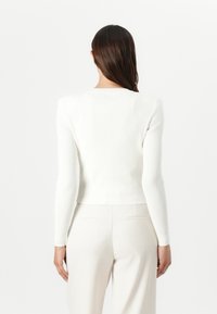 White ribbed sweater with long sleeves and a cropped design, featuring a round neckline and a soft texture. Worn with light-colored pants.