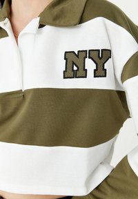 Polo de ganchillo en verde oliva con rayas blancas, que presenta un bordado de "NY" en verde oscuro. Tejido de algodón suave, diseño corto.