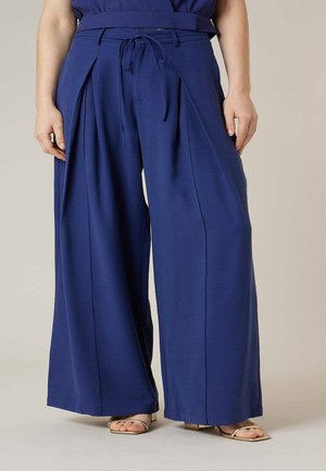 Femme portant un pantalon taille haute, large et bleu marine avec ceinture et détail noué, associé à des sandales ouvertes dorées.