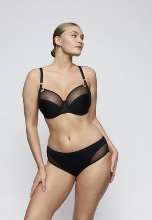 Woman wearing a black bra with mesh details and matching high-cut panties, standing with one hand on her hip, looking to the side.